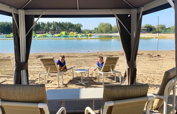 Two kids smile for the camera while relaxing on beach chairs outside their private cabana at Bear Paw Adventure Park on a warm sunny day.