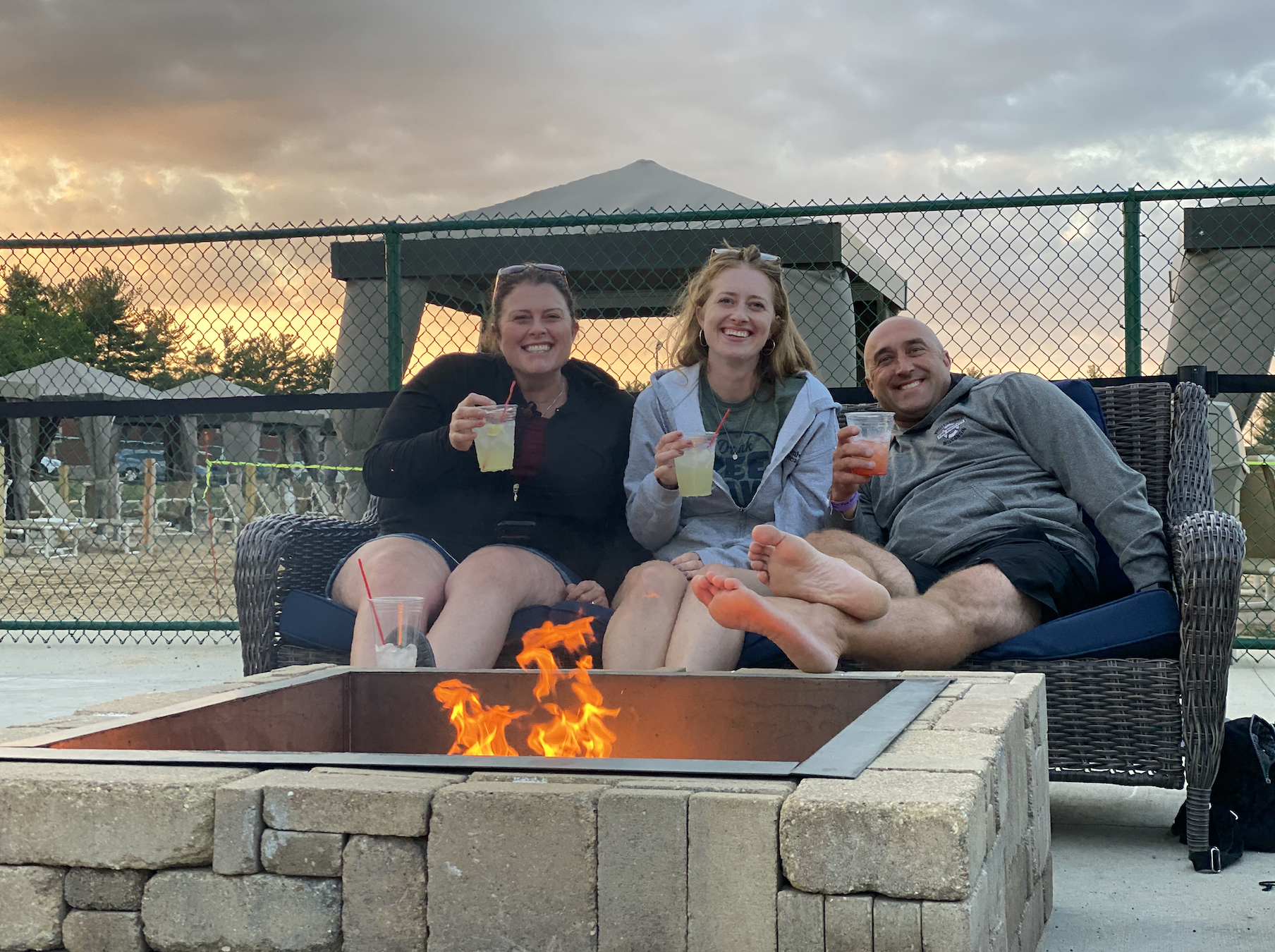 Three adults smile for the camera while they are enjoying drinks by a fire at Bear Paw Adventure Park.